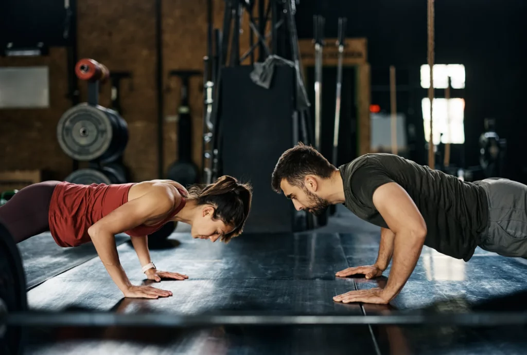 A man and a woman facing each other as they perform synchronized push-ups in a gym, showing focus and determination.