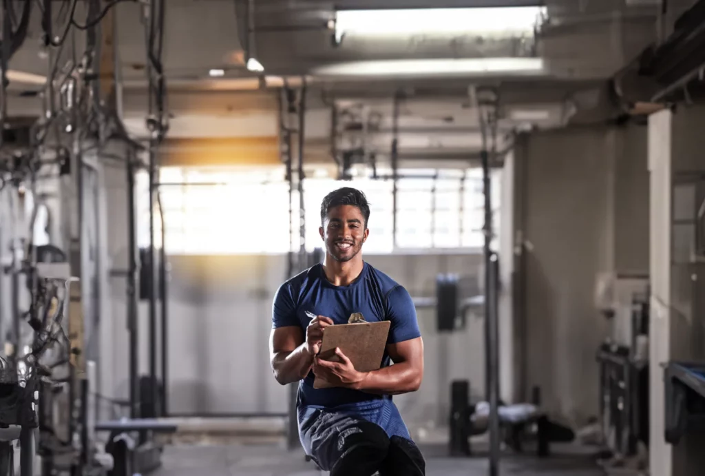 A male personal trainer wearing a blue shirt smiles while holding a clipboard and pen at the gym.