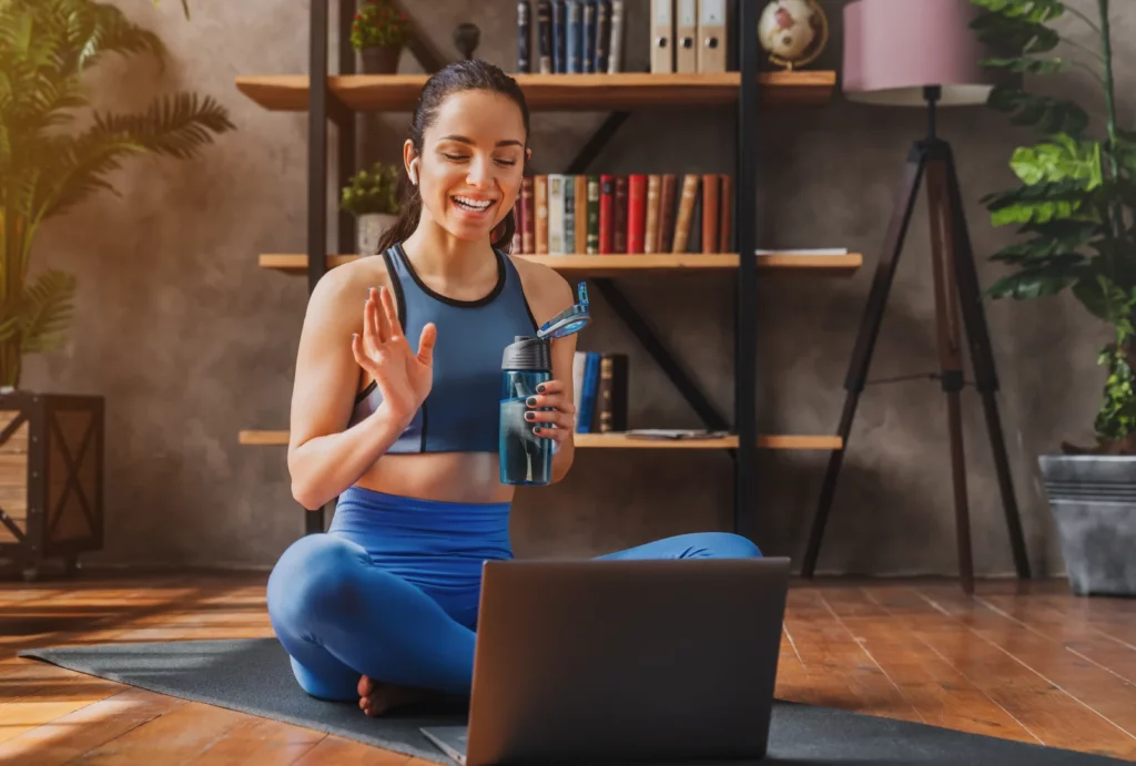 A woman wearing a blue sports bra and blue leggings sits cross-legged on a sports mat while waving at her laptop camera and holding a clear blue water bottle.