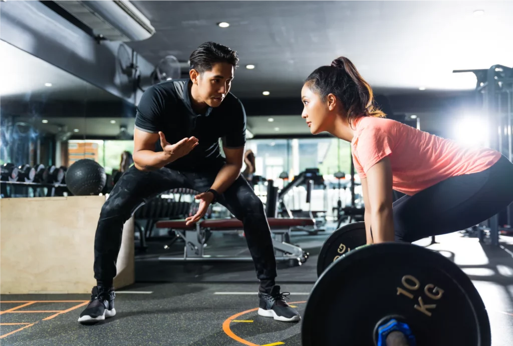 A male personal trainer wearing a black outfit motivates a client wearing a pink shirt and black leggings as she lifts weights at the gym.