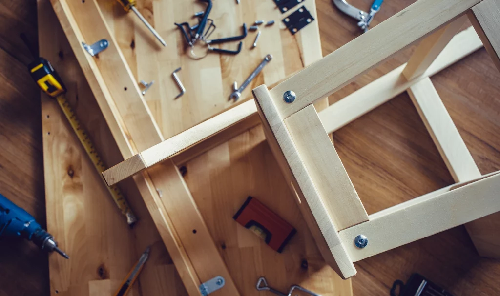 An overhead shot of a wooden chair being constructed by hand. It is lying on a wood table and is surrounded by woodworking tools.