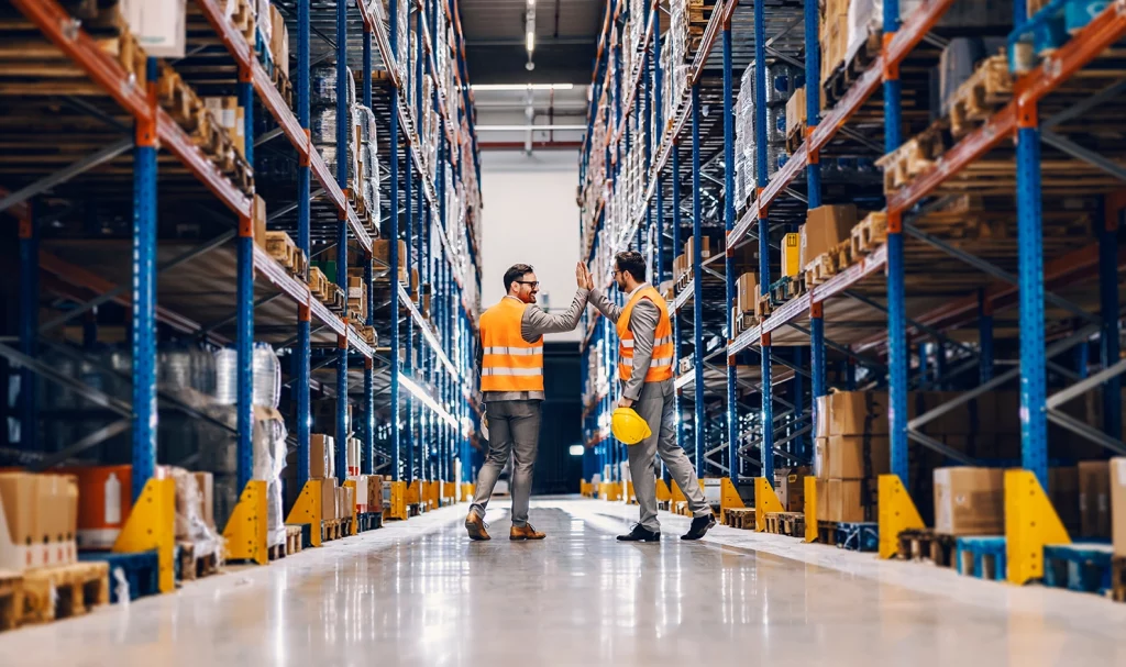 Two warehouse workers high five one another while they are in their high visibility workwear and holding their hard hats, and standing in between large industrial shelves holding products.