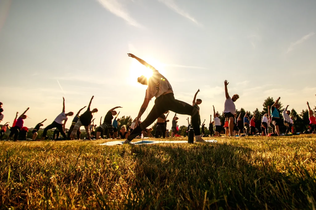 A large outdoor yoga class with people stretching in warrior pose as the sun sets, creating a serene atmosphere in an open field.