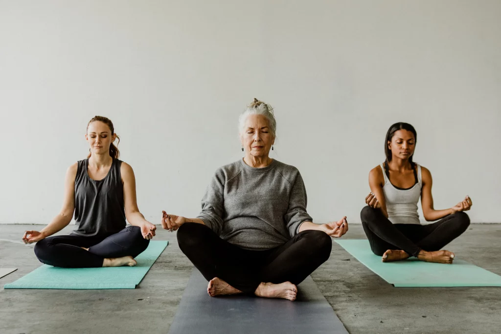 Three women of different ages sitting in a meditative posture on yoga mats, focusing on mindfulness in a minimalistic studio.