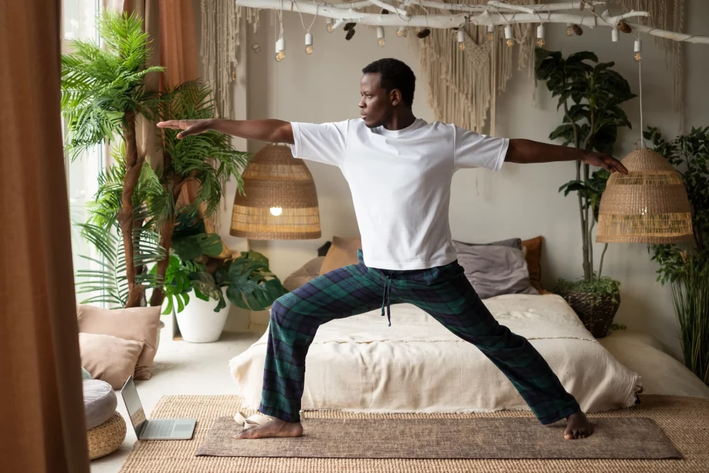 A man practicing warrior pose in a cozy, bohemian-styled room filled with plants and warm-toned decor, exuding calm and relaxation.