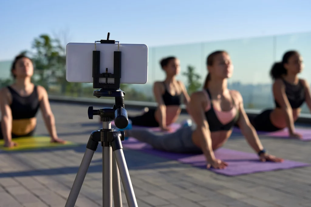 A group of women participating in a yoga class, filmed with a smartphone on a tripod. The focus is on the filming setup, with the women in the background doing cobra pose on their yoga mats.