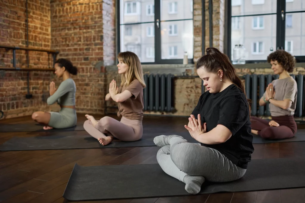 Four women sitting cross-legged on yoga mats, meditating in a loft-style yoga studio with exposed brick walls and large windows.