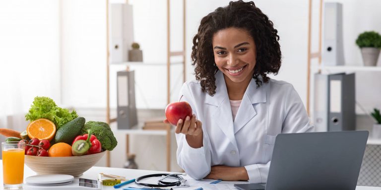 A nutritionist in a white lab coat holds an apple. a bowl of fruits and vegetables sit on her desk.