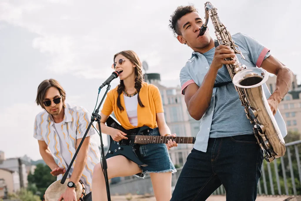 Trio of musicians including a man in a light blue shirt and dark jeans playing a saxophone in the front, a woman in denim shorts and a yellow cardigan playing a guitar and singing in the middle, and a man wearing sunglasses and a white shirt playing a drum in the back.