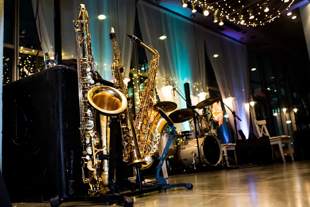 A nighttime shot of a series of saxophones on stands and a drum kit on an empty level stage featuring large windows with white drapes in the background.