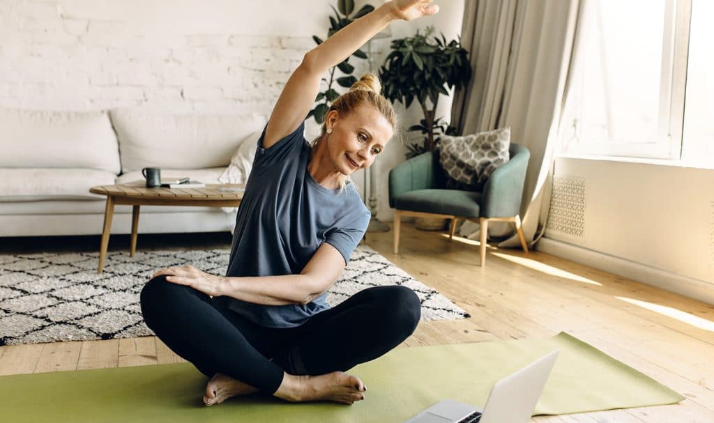 An older woman sits on her yoga mat in her home and stretches as her virtual yoga teacher is broadcasting a class on the laptop.