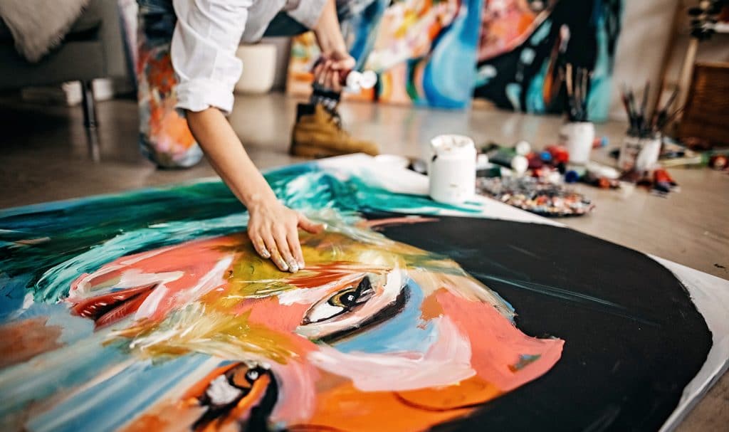 Woman uses hands to paint portrait on a canvas on the floor of her studio.