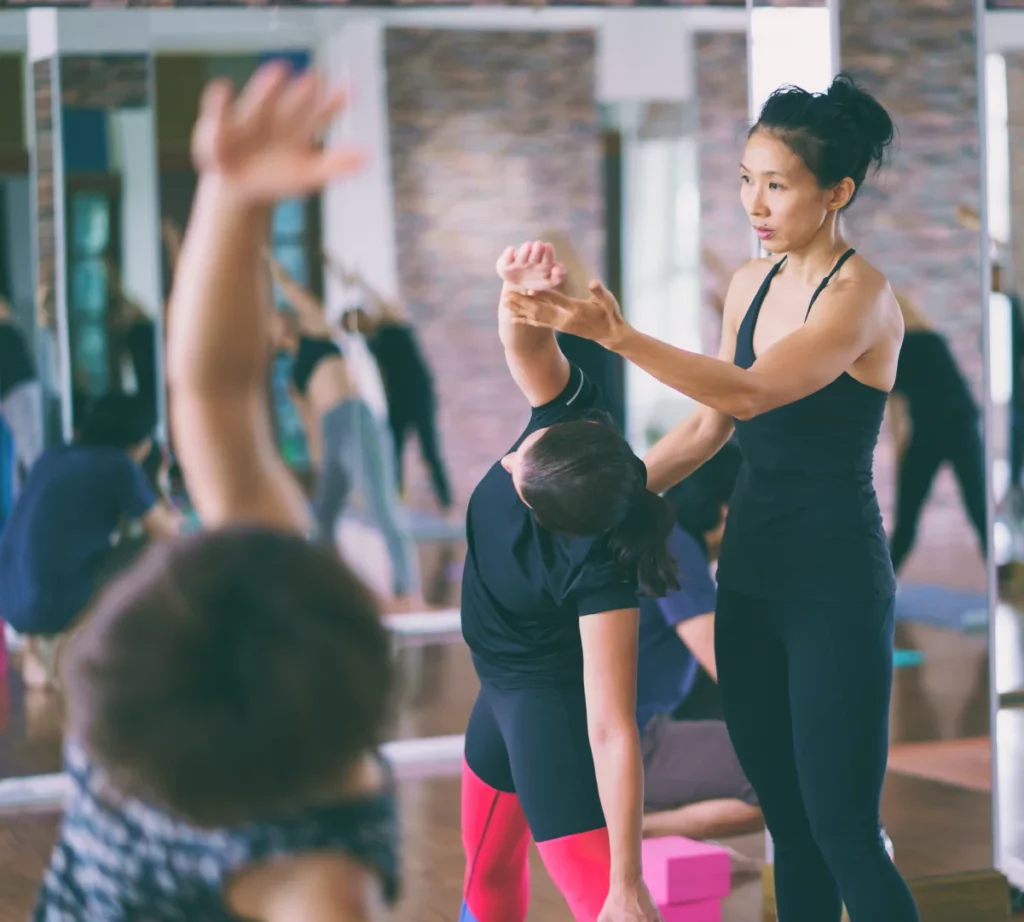 Yoga instructor with dark hair, wearing a black tank top and leggings helps adjust a student during a class.