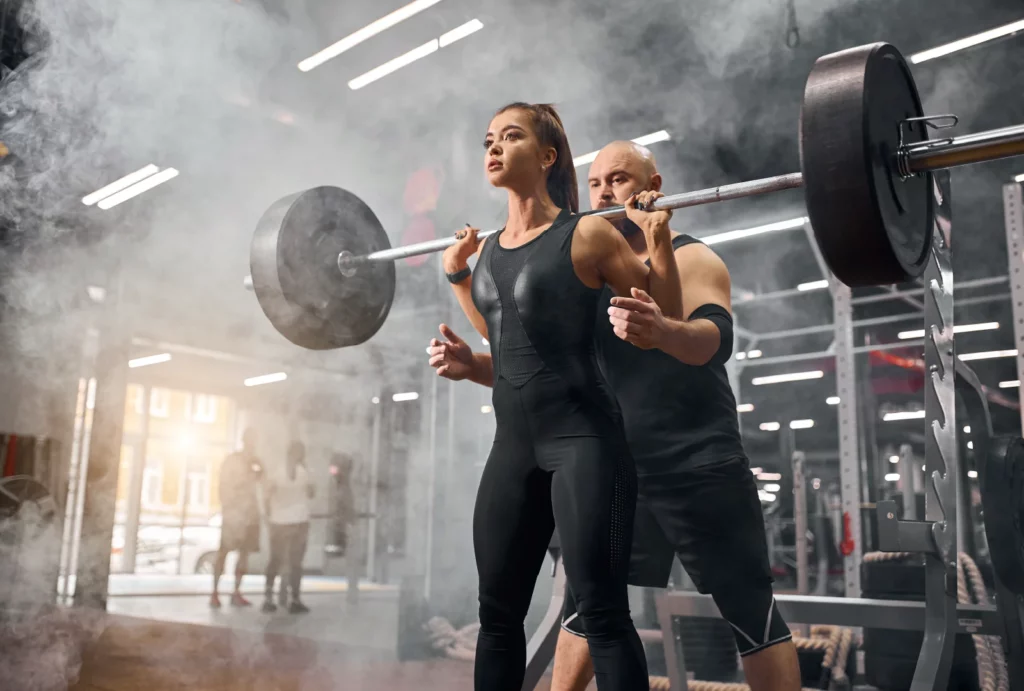 A female athlete lifts a barbell while her trainer provides guidance and support in a gym, surrounded by gym equipment and a misty atmosphere.