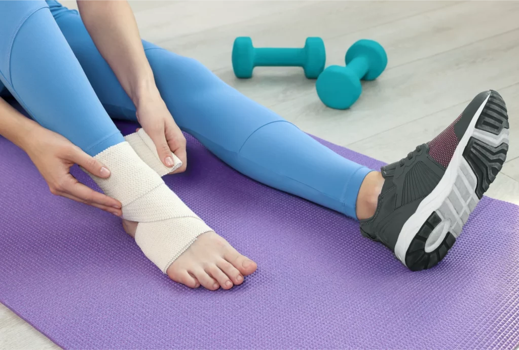 Yoga student in blue leggings sitting on a purple mat wrapping a bandage around a sprained ankle.