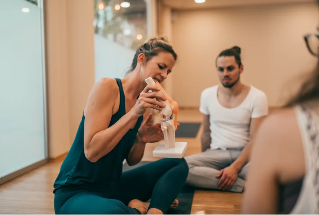 A blonde yoga instructor wearing dark green athletic clothes explaining the anatomy of a knee joint with a prop to a class.