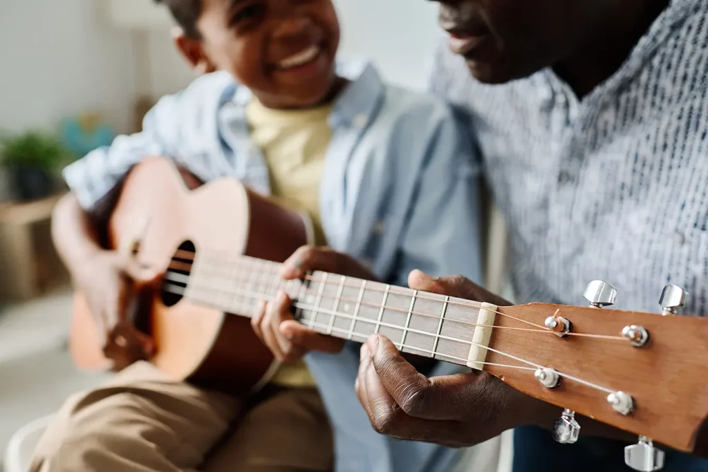 A close-up of a child smiling while learning to play guitar, as an adult guides their fingers on the strings.