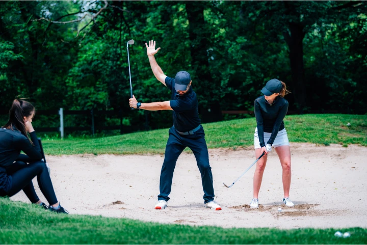Golf instructor demonstrates a bunker shot to a student on a golf course.