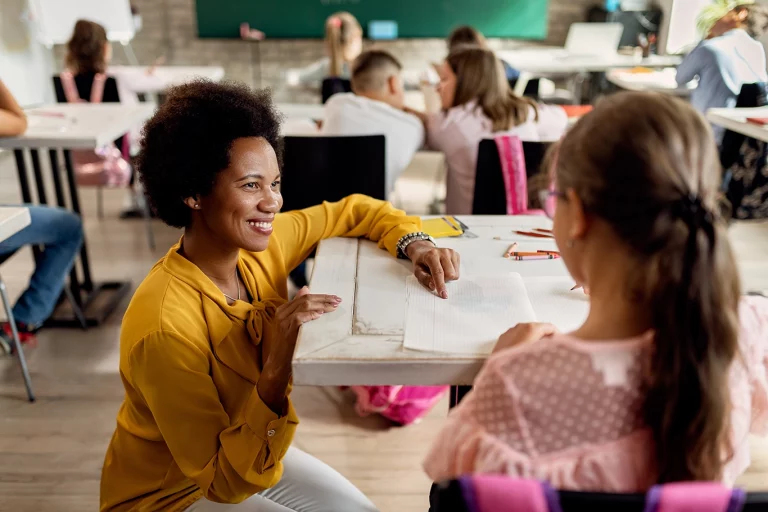 A teacher kneeling down at her students desk, pointing to a piece of paper and smiling.