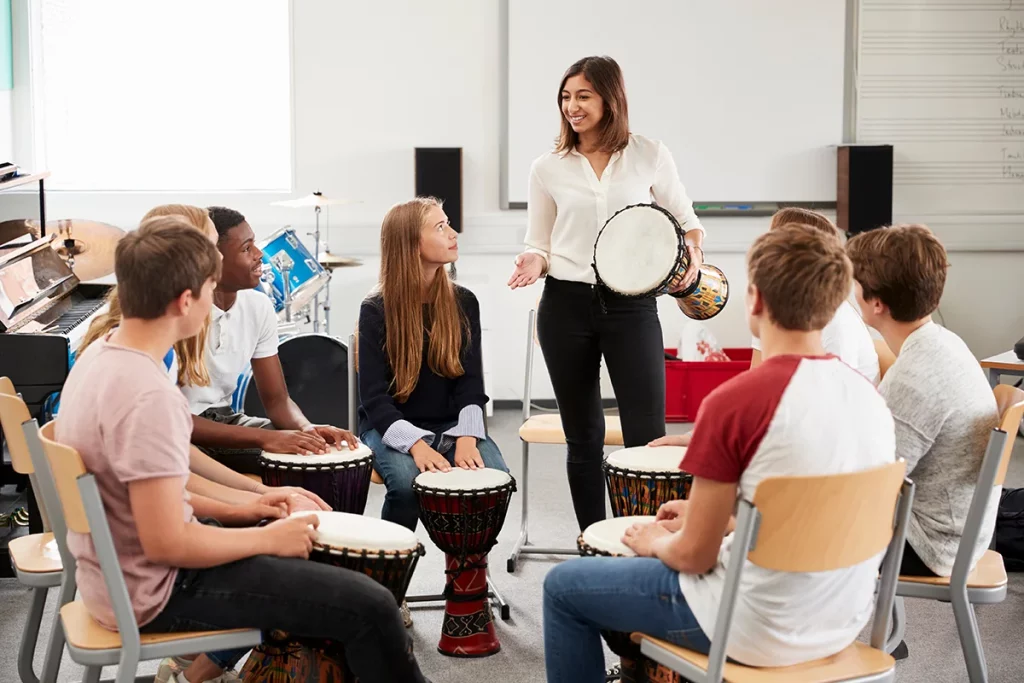 A woman standing in front of a circle of students giving a lesson on how to play the djembe drum.