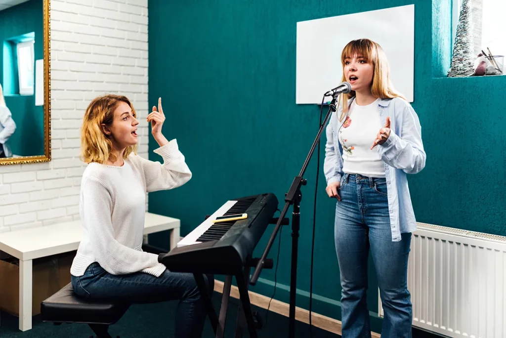 A woman receives vocal instruction from a teacher sitting at a keyboard.