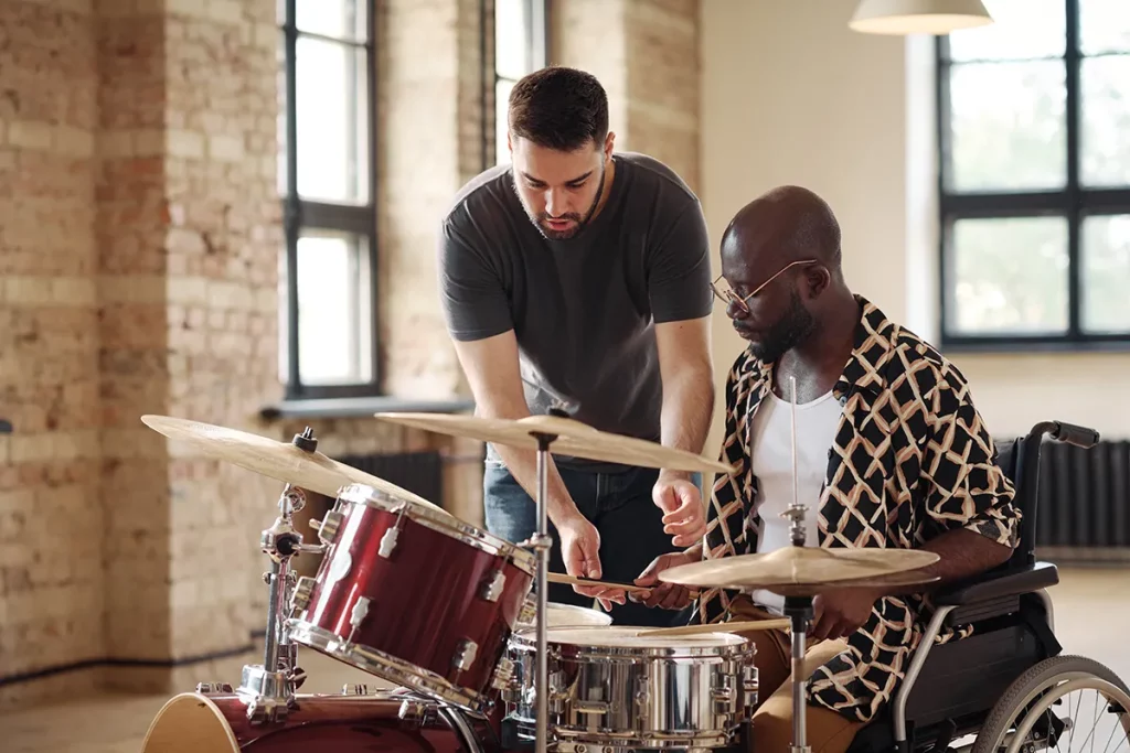 A music teacher teaching a man in a wheel chair how to play the drums.