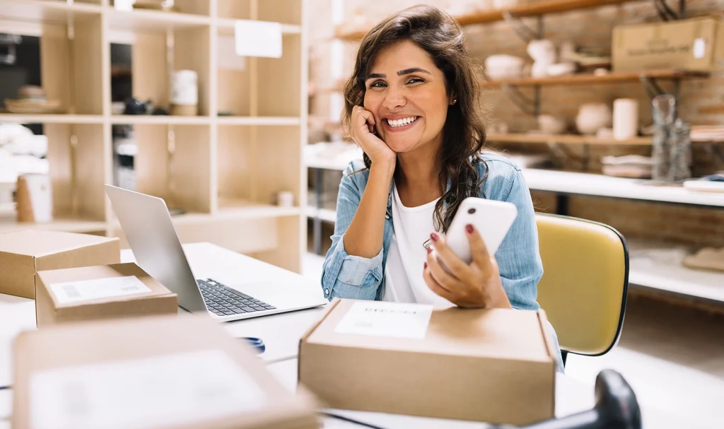 A female online seller is smiling next to a package as she holds her phone and works on her laptop in her home office.