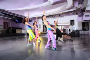 Four Zumba dancers wearing colorful outfits practice a move in a tiled studio with audio and lighting equipment.