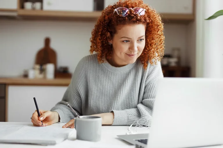 A woman with curly hair, sitting at a desk in front of her laptop taking notes.