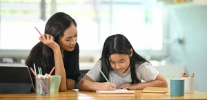 A woman tutors a young girl at home as she writes something on a notebook.