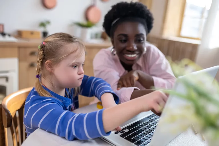 A young girl with Down Syndrome pointing to a laptop screen while receiving a lesson from her teacher.