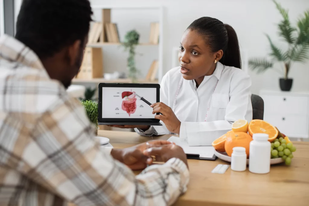 Dietician with black hair, wearing a white coat, seated at a desk next to a plate of fruit and displaying an image of the gastrointestinal tract on a tablet to a client.