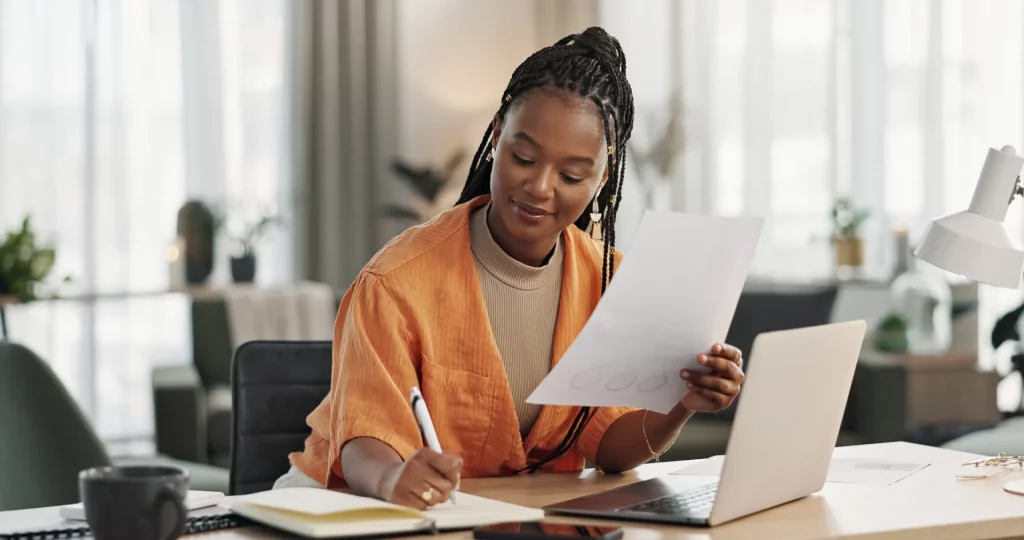 Woman with black braided hair wearing an orange shirt, seated at a desk, holding a piece of paper and taking notes with an open white laptop in front of her.