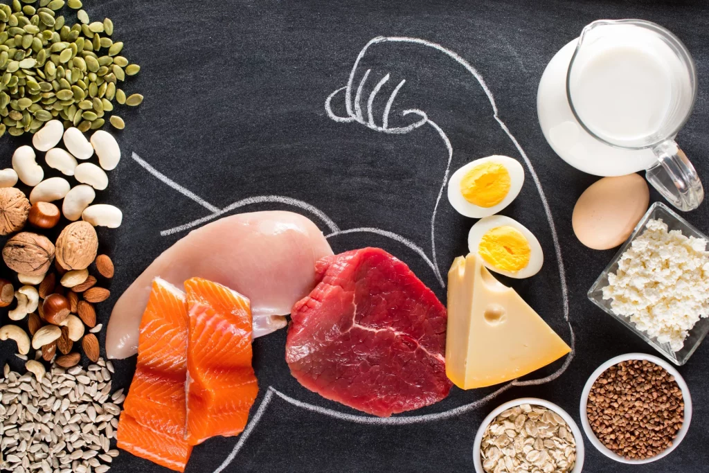 A black chalkboard featuring the outline of a flexing arm drawn in white chalk, with various meats, nuts, and dairy products surrounding and filling in the drawing.