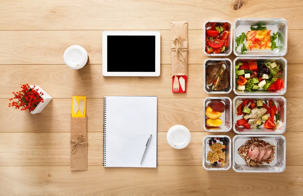 A birds eye view of a light brown table featuring a blank white notepad, a tablet with a black screen, white disposable coffee cups, brown disposable cutlery, and a series of eight rectangular containers filled with various fruits, vegetables, and other food.