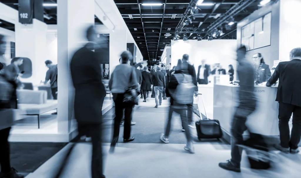 A black and white shot of event-goers walking amongst the aisle of booths at a tradeshow.