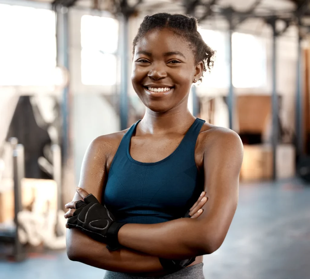 A young female CrossFit coach wearing a blue sports bra and black gloves crosses her arms and smiles at the gym.