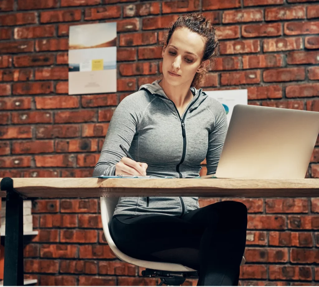 A female CrossFit coach wearing a grey hoodie writes notes on a notepad while sitting in front of her laptop at the gym.