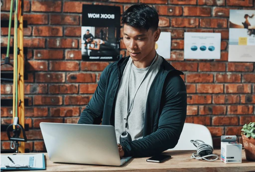 A male CrossFit coach wearing a grey t-shirt and dark blue jacket works on his laptop at his gym desk in front of a brick wall.