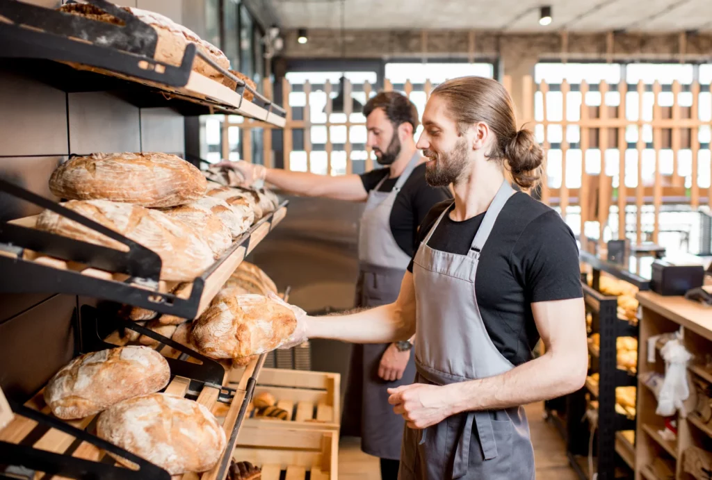 Two male bakers in aprons selecting fresh bread loaves from shelves in a bakery shop.