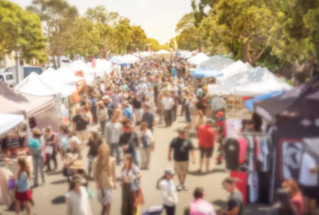 A vibrant and busy outdoor market filled with people browsing vendor booths on a sunny day.