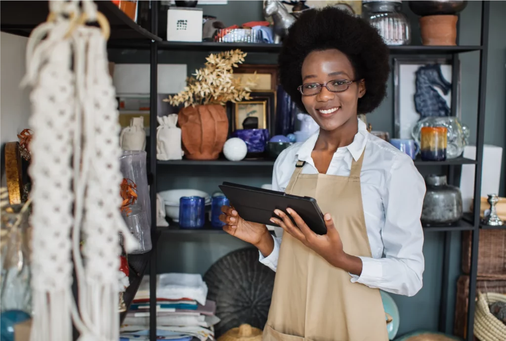 A female craft shop owner smiling while using a tablet in her shop, surrounded by handmade goods and decorative items.