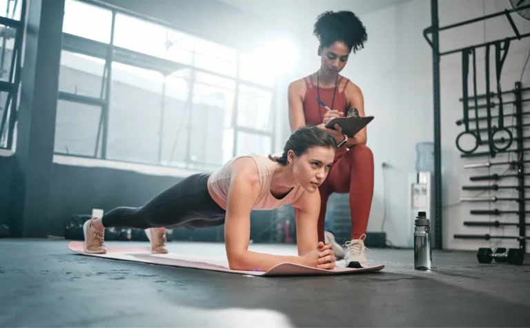 A young woman wearing a tank top and leggings performs a plank on a mat while her trainer wearing a red workout set takes notes on a clipboard.