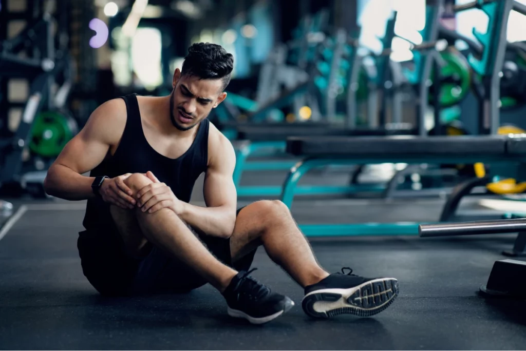 A man wearing a black tank top and shorts holds his injured knee and grimaces while sitting on a gym floor.