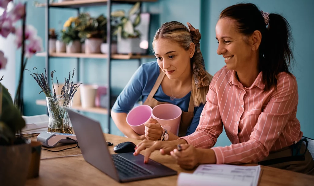 Two female florists in their shop using a laptop on a wooden table top workspace as they look at vendor insurance online for an upcoming event.