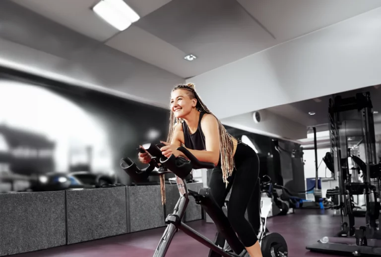 A woman with long braids smiling as she rides a stationary spin bike in a modern gym, conveying enjoyment and motivation during her workout.