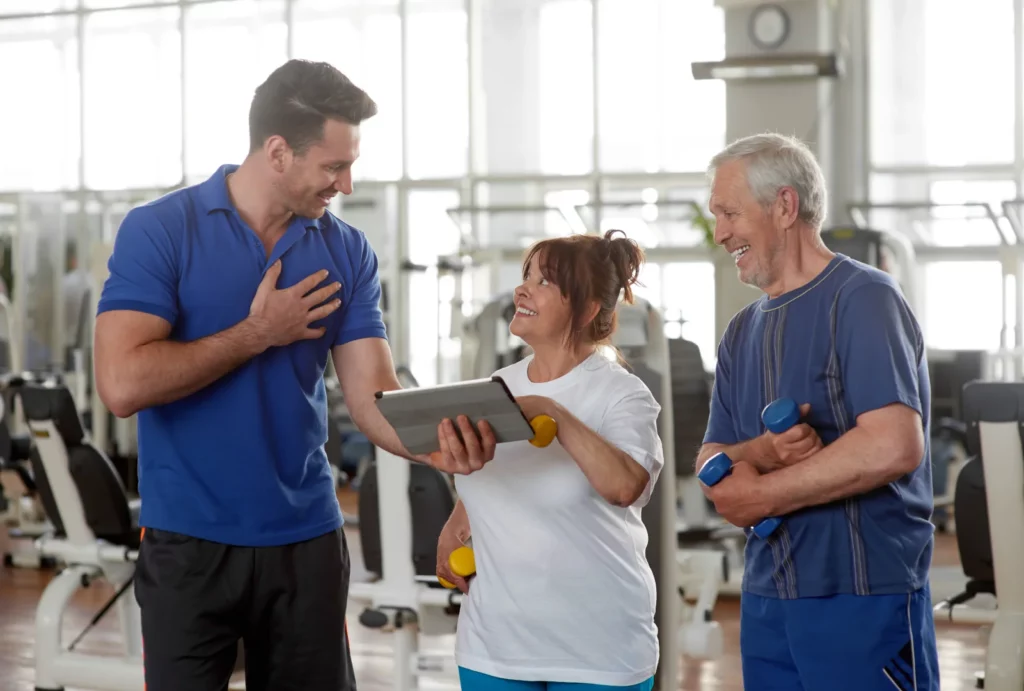 Personal male trainer showing something on digital tablet. Happy senior couple at gym.