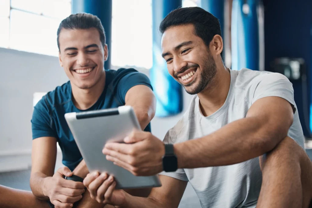 A male personal trainer wearing a gray shirt smiles and holds a tablet to show his client, another man wearing a dark blue shirt, while they sit on the floor of a gym.