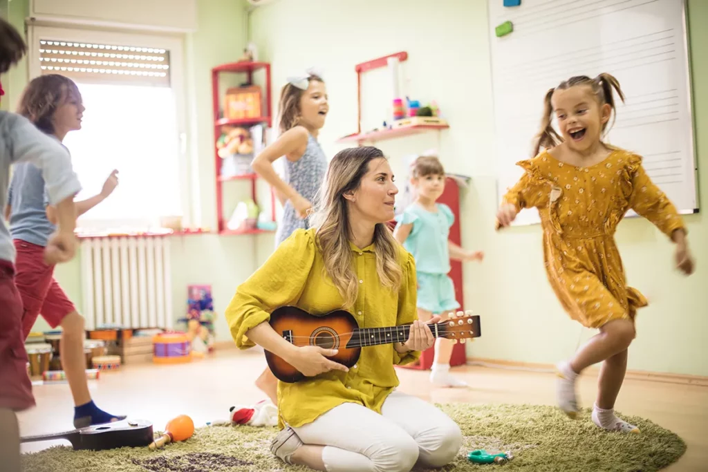 A teacher plays the ukulele while young children dance to the music.