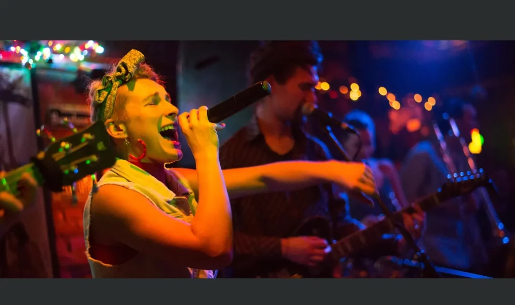 Band performing on stage with lead singer in focus, wearing a denim vest and red hoop earrings whiling singing into a microphone.